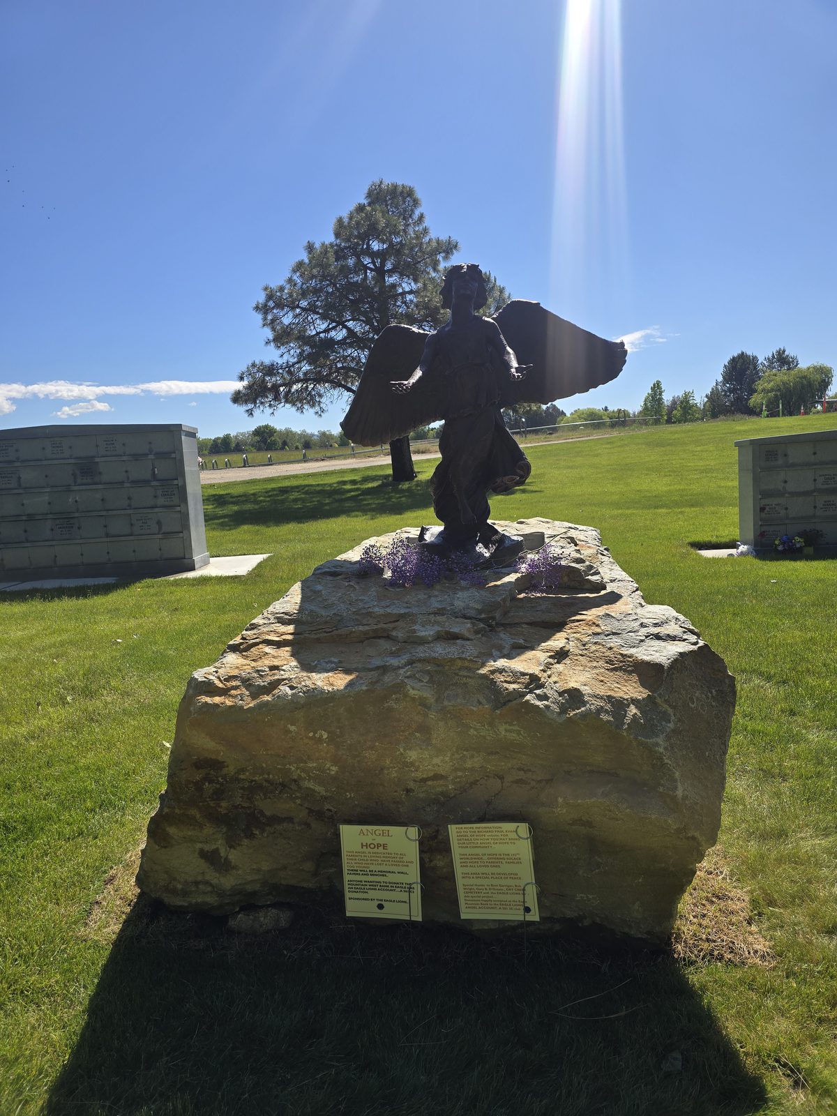 The Angel of Hope statue standing on its stone base at Dry Creek Cemetery, with the memorial walls behind