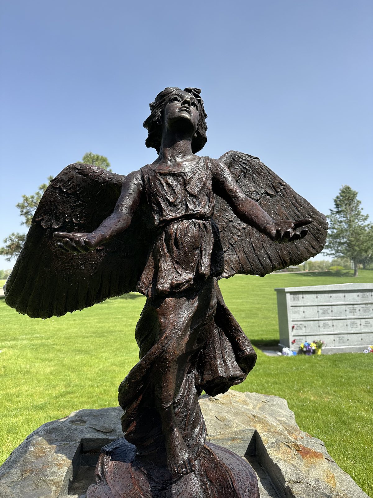 A close view of the Angel of Hope statue, wings raised and face turned upward, the bronze still wet with morning dew